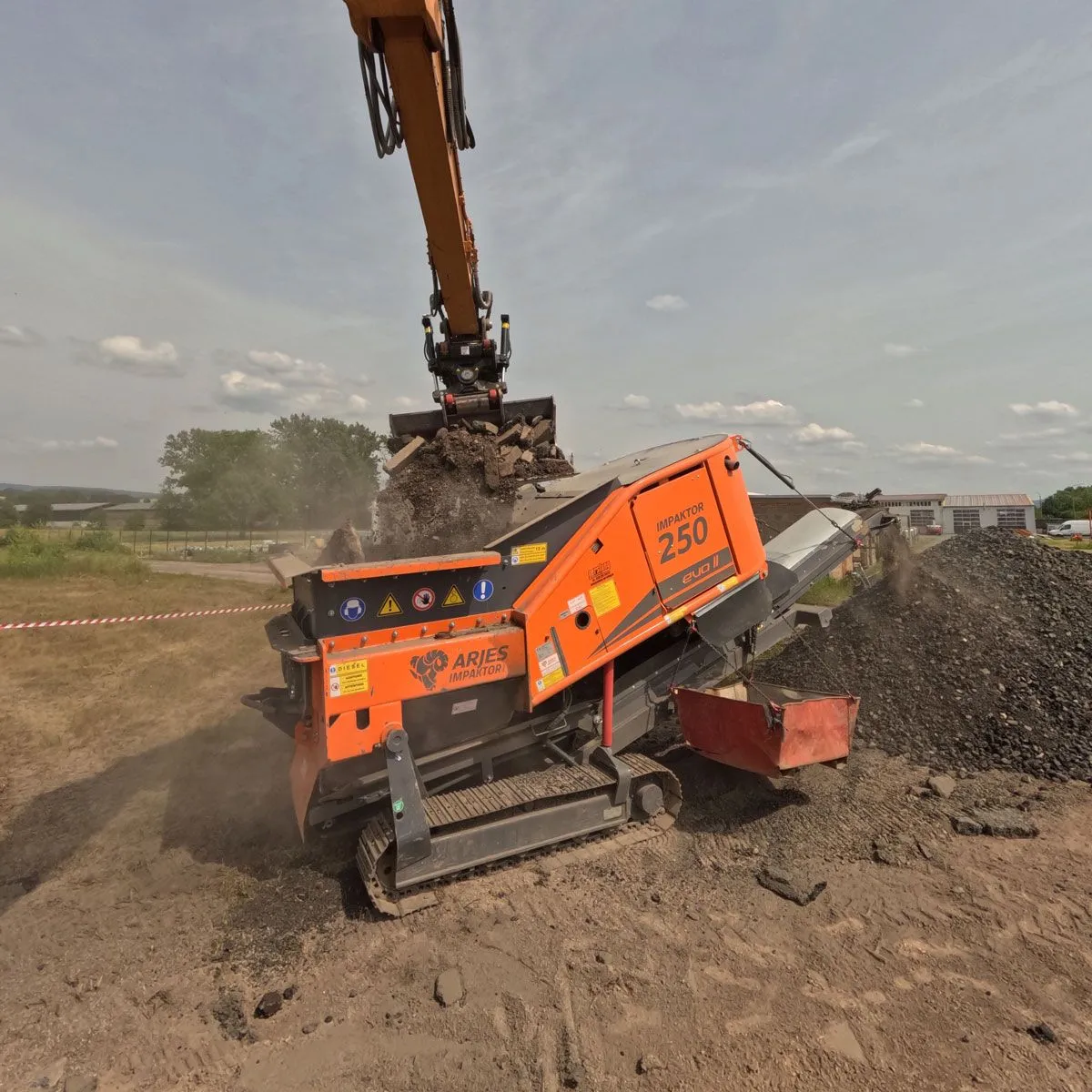Bagr nakládá vybouraný materiál do drtiče ARJES IMPAKTOR 250 EVO II při drcení na stavbě.